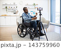 A wheelchair user reviews printed business charts while recording himself on a smartphone tripod in a bright living room with modern furnishings 143759960