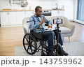A wheelchair user gestures toward his laptop while recording a video on a smartphone, surrounded by a bright kitchen and comfortable living space 143759962