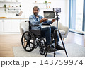 A wheelchair user presents a tablet toward a mounted smartphone in a bright home, creating accessible online content from a comfortable living space 143759974