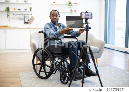 A wheelchair user presents a tablet toward a mounted smartphone in a bright home, creating accessible online content from a comfortable living space 143759974