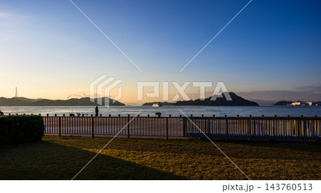 早朝の港を望む海辺の芝生の広場と穏やかな青い海、島々の稜線と朝焼けの空に浮かぶ船、散策する人物の風景 早朝の港を望む海辺の芝生の広場と穏やかな青い海、島々の稜線と朝焼けの空に浮かぶ船、散策する人物の風景 143760513