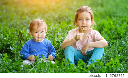 Smiling Toddler Sits Beside Young Sister, Relaxing Safely Upon Sunny Green Park Lawn. Thoughtful Girl Holding Delicate Dandelion Flower Enjoys Peaceful Family Recreation Within Garden Grounds. 143762755