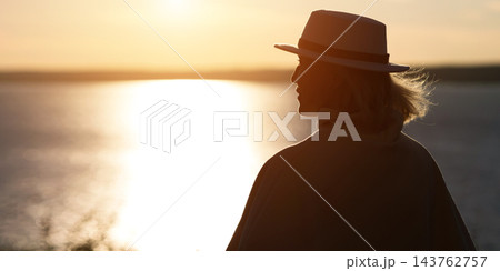 Woman stands quietly beside glowing water, wearing fedora and looking toward sunset. Dark silhouette, relaxed posture, and warm reflected sunlight create serene contemplative mood. 143762757
