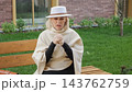Woman seated on wooden bench holds clasped hands near her face in outdoor contemplation. Her thoughtful expression and cream hat add refined style beside brick building and green lawn. 143762759