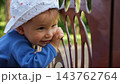 Joyful Infant Smiles Near Metal Railing In Tranquil Summer Garden Setting. Curious Little Child In Patterned Sun Hat Gazes Away, Showing Pure Happiness Amid Blurred Trees And Sunlight Outdoors. 143762764