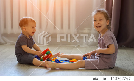 Joyful Siblings Assemble Colorful Toy Bricks Across Living Room Floor, Giggling Cheerfully During Leisure Time While Scattered Small Construction Pieces Represent Possible Home Safety Hazard. Joyful Siblings Assemble Colorful Toy Bricks Across Living Room Floor, Giggling Cheerfully During Leisure Time While Scattered Small Construction Pieces Represent Possible Home Safety Hazard. 143762767