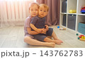 Smiling Girl Holds Little Brother Closely Near Sunlit Window In Cozy Playroom. Tender Sister Supports Barefoot Toddler Sibling With Gentle Care, Expressing Innocent Love During Play Session. 143762783