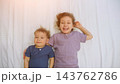 Two Cheerful Siblings Rest Together On White Bed, Smiling Joyfully During Bedtime Routine; Happy Girl Embraces Younger Toddler Brother While Staying Awake Under Bright Bedroom Illumination. 143762786