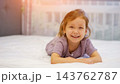 Happy Young Girl Rests Peacefully On Comfortable Bed Inside Cozy Modern Bedroom. Delighted Child Smiles Toward Camera While Relaxing On Clean White Sheets During Pleasant Sunny Morning At Home. 143762787