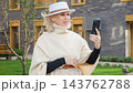Woman poses with smartphone in outdoor portrait near modern building. She wears white hat and cream cape, and her smiling expression gives personal profile image stylish, elegant feel. 143762788