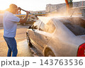 Warm sunset light view of a man rinsing soapy foam off silver car with high pressure water jet 143763536