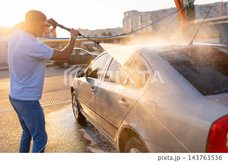 Warm sunset light view of a man rinsing soapy foam off silver car with high pressure water jet 143763536