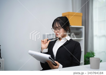Thoughtful young woman holding pen to mouth while reading document in bright office with modern decor 143764184