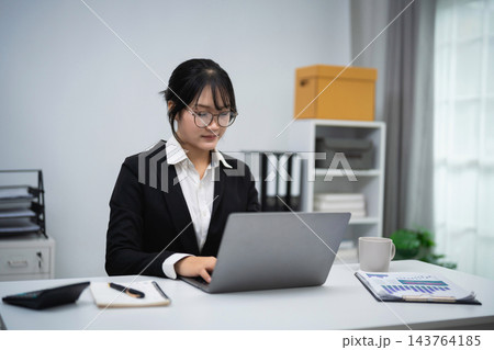 Young professional woman in black suit working focused on laptop at modern office desk with documents and coffee cup 143764185