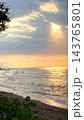 Tropical beach rests beneath radiant sunset, with two people standing in water. Warm reflections brighten waves as distant boats and framing greenery complete serene vacation setting. 143765801