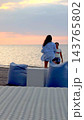 Couple smiles together beside sea at sunset, with woman facing her partner on sandy shore. Beachside pair enjoy relaxed romantic moment near blue lounge cushions and wicker furniture. 143765802