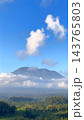 Volcanic mountain stands above forest and farmland, partly veiled by low clouds. Bright cumulus clouds float across blue sky, giving this tranquil tropical landscape dramatic scale. 143765803