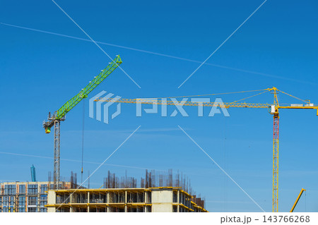Construction Cranes at Building Site against Blue Sky. High-rise building under construction with green and yellow tower cranes. Industrial background Construction Cranes at Building Site against Blue Sky. High-rise building under construction with green and yellow tower cranes. Industrial background 143766268