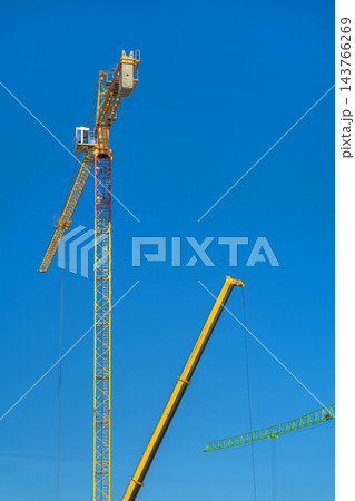 Tower Crane, Mobile Crane Against Blue Sky, Construction Site. Tower crane rises above construction site with mobile crane, suspended lifting cables. Industrial engineering, heavy lifting equipment Tower Crane, Mobile Crane Against Blue Sky, Construction Site. Tower crane rises above construction site with mobile crane, suspended lifting cables. Industrial engineering, heavy lifting equipment 143766269