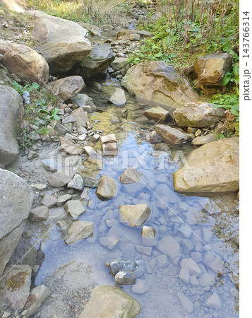 Clear shallow mountain stream flowing between large boulders and scattered stones, with blue sky reflections and green plants on the banks Clear shallow mountain stream flowing between large boulders and scattered stones, with blue sky reflections and green plants on the banks 143766314