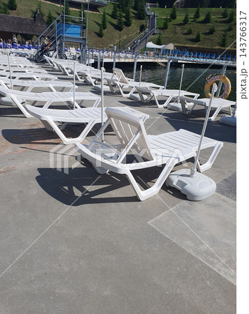 Rows of empty white plastic sun loungers with umbrella bases on a concrete lakeside pier under bright sunshine. Summer resort beach, vacation and relaxation Rows of empty white plastic sun loungers with umbrella bases on a concrete lakeside pier under bright sunshine. Summer resort beach, vacation and relaxation 143766317