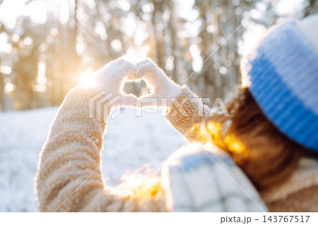 Woman in hat and scarf against backdrop of snowy winter forest holds hands in the shape of a heart. 143767517