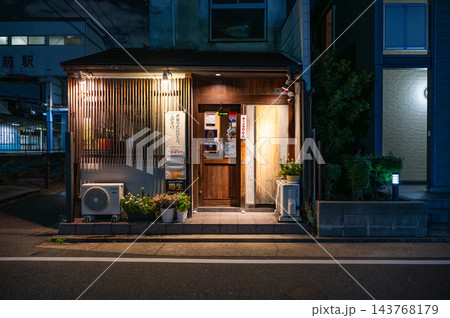 夜の路地に暖色の灯りがともる木製格子と木の扉の飲食店、鉢植えと室外機が並ぶ日本の小さな店舗外観の夜景 夜の路地に暖色の灯りがともる木製格子と木の扉の飲食店、鉢植えと室外機が並ぶ日本の小さな店舗外観の夜景 143768179