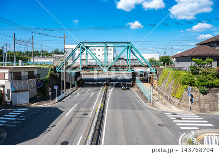 青空の下で道路をまたぐ鮮やかな水色の鉄道トラス橋と架線設備、車道や横断歩道が広がる日本の住宅街の風景 青空の下で道路をまたぐ鮮やかな水色の鉄道トラス橋と架線設備、車道や横断歩道が広がる日本の住宅街の風景 143768704