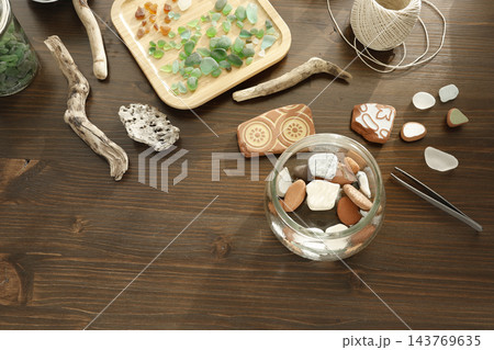 Glass jar with smooth beach pebbles and sea pottery shards on dark wood table. Flatlay coastal treasures with driftwood twine cord tweezers. Sustainable zero waste ocean DIY home decor project. 143769635