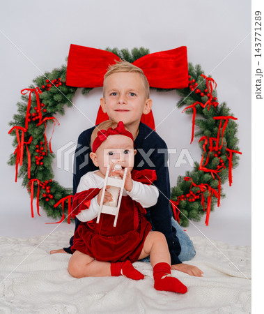 Older brother sitting behind his baby sister holding small wooden rocking horse during Christmas studio photo shoot. Christmas wreath with red bow decoration, sibling love, and holiday season 143771289