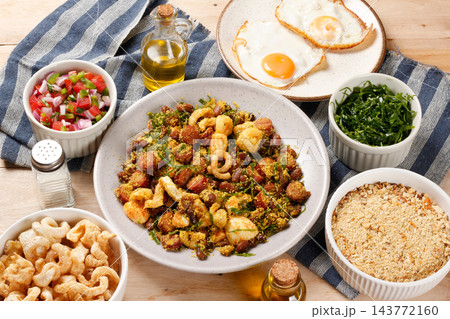 Traditional Brazilian feijao tropeiro served on a rustic ceramic plate, accompanied by eggs, pork rinds and a fresh tomato vinaigrette. Traditional Brazilian feijao tropeiro served on a rustic ceramic plate, accompanied by eggs, pork rinds and a fresh tomato vinaigrette. 143772160