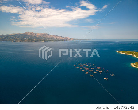 Fish farming pens float in open coastal water, arranged in compact offshore cluster. Net cages and small boats mark aquaculture site, with islands and mountains along horizon, enclosing fish. Fish farming pens float in open coastal water, arranged in compact offshore cluster. Net cages and small boats mark aquaculture site, with islands and mountains along horizon, enclosing fish. 143772342