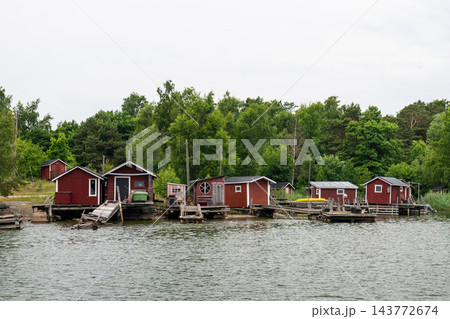 Traditional red wooden boathouses along peaceful Nordic shoreline with rustic fishing piers and lush forest backdrop Traditional red wooden boathouses along peaceful Nordic shoreline with rustic fishing piers and lush forest backdrop 143772674