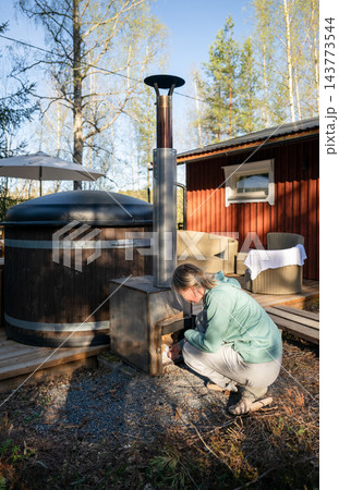 Woman heating traditional outdoor hot tub with wood stove at cozy cabin in Finnish forest Woman heating traditional outdoor hot tub with wood stove at cozy cabin in Finnish forest 143773544