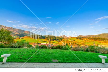 Green grass field on hill. Landscape of village in valley. Pine forest on the mountain with blue sky and white clouds on sunny day in Europe. Empty seat in the garden at the park. Small city in valley Green grass field on hill. Landscape of village in valley. Pine forest on the mountain with blue sky and white clouds on sunny day in Europe. Empty seat in the garden at the park. Small city in valley 143774539