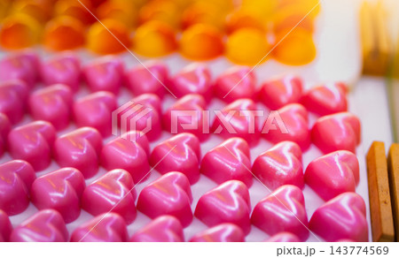 Pink heart shaped chocolate pralines with rose scented white chocolate ganache on a white plate, with yellow and orange dome chocolates nearby, a luxury confectionery assortment for Valentine's Day. Pink heart shaped chocolate pralines with rose scented white chocolate ganache on a white plate, with yellow and orange dome chocolates nearby, a luxury confectionery assortment for Valentine's Day. 143774569
