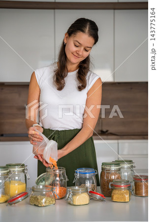 Smiling woman pours red lentils into a glass jar among assorted grains in a modern kitchen. Pantry organization with reusable containers supports sustainable living and reduces packaging waste Smiling woman pours red lentils into a glass jar among assorted grains in a modern kitchen. Pantry organization with reusable containers supports sustainable living and reduces packaging waste 143775164