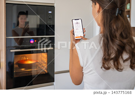 Woman uses a smartphone app to control a built-in oven in a modern home kitchen. Smart home technology simplifies everyday cooking through wireless appliance management Woman uses a smartphone app to control a built-in oven in a modern home kitchen. Smart home technology simplifies everyday cooking through wireless appliance management 143775168