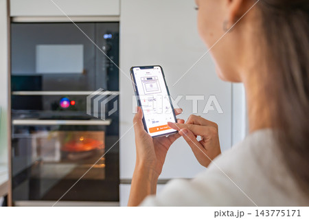 Woman starts a smart oven by tapping a control button on her smartphone in a modern kitchen. Connected home technology enables remote cooking management through a mobile app 143775171