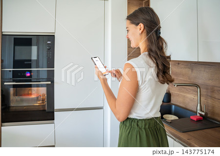 Woman uses a smartphone app to control a built-in oven in a modern home kitchen. Smart home technology simplifies everyday cooking through wireless appliance management Woman uses a smartphone app to control a built-in oven in a modern home kitchen. Smart home technology simplifies everyday cooking through wireless appliance management 143775174