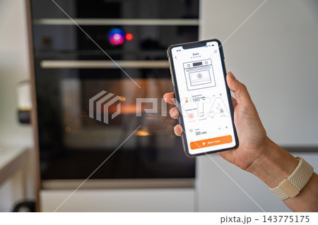 Hand holds a smartphone displaying a smart oven control app with temperature, timer and heating settings. Remote appliance management illustrates IoT technology and home automation Hand holds a smartphone displaying a smart oven control app with temperature, timer and heating settings. Remote appliance management illustrates IoT technology and home automation 143775175