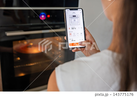 Woman controls a smart oven using a mobile app on her smartphone in the kitchen. Remote temperature and timer settings demonstrate smart home automation and connected appliances 143775176