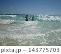 two people standing and playing with waves on a beach, joyful and relaxed in a serene ocean setting, from the shore looking out to sea, with clear blue sky and sandy shore. two people standing and playing with waves on a beach, joyful and relaxed in a serene ocean setting, from the shore looking out to sea, with clear blue sky and sandy shore. 143775703