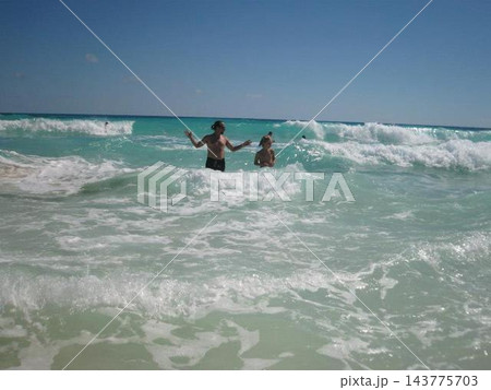 two people standing and playing with waves on a beach, joyful and relaxed in a serene ocean setting, from the shore looking out to sea, with clear blue sky and sandy shore. two people standing and playing with waves on a beach, joyful and relaxed in a serene ocean setting, from the shore looking out to sea, with clear blue sky and sandy shore. 143775703