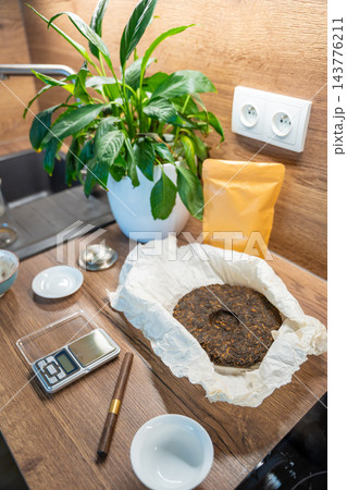 A compressed puer tea cake lies on paper beside a digital scale and tea pick. Measuring loose portions prepares Chinese tea for home brewing. 143776211