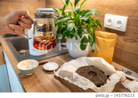 A glass teapot of amber puer tea is held above a kitchen counter. The finished citrus infused brew is separated from the leaves for serving. 143776213
