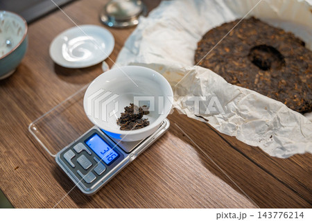 A portion of puer tea rests in a white cup on a digital scale beside a tea cake. Weighing leaves establishes the dose for brewing Chinese tea. A portion of puer tea rests in a white cup on a digital scale beside a tea cake. Weighing leaves establishes the dose for brewing Chinese tea. 143776214