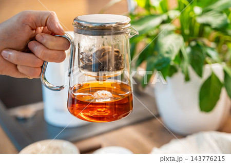 A hand holds a glass teapot with amber puer tea beneath a leaf filled infuser. Brewing with aged mandarin peel prepares an aromatic tea for drinking. A hand holds a glass teapot with amber puer tea beneath a leaf filled infuser. Brewing with aged mandarin peel prepares an aromatic tea for drinking. 143776215