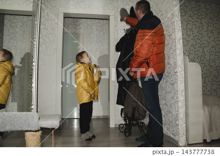 A child in a yellow jacket returns from school with their dad, takes off their outerwear, and goes inside their home. 143777738
