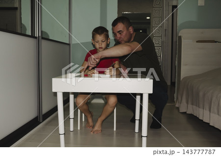A child returns from school wearing red T shirt and sits down white table to do their schoolwork and homework with help of their father. A child returns from school wearing red T shirt and sits down white table to do their schoolwork and homework with help of their father. 143777787